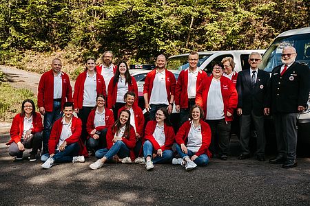 Die Helfer:innen in weißen Polos und roten Fleecejacken sitzend und stehend als Gruppe. Alle lachen und sehen glücklich aus.
